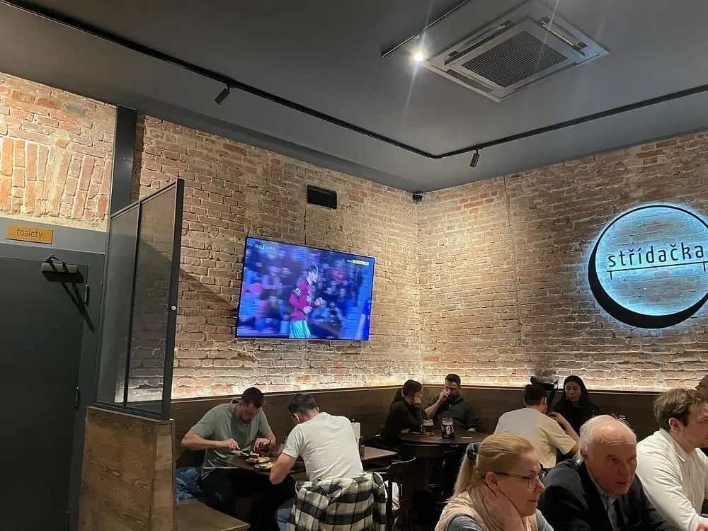 interiér restaurace stridacka, kde v televizi je fotbal a ve spodu fotografie je vidět jak zákaznící sedí u stolu a jedí