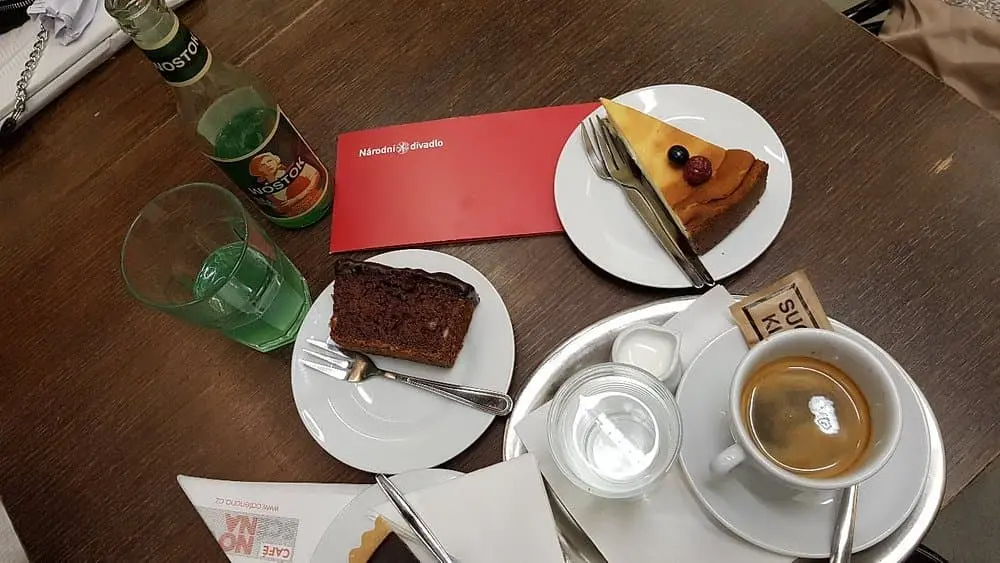 na stole máme 3 talířky na levém je brownies, na hornim je koláč, a na pravém je kafe s cukrem. Taktéž tu je sklenice a láhev 