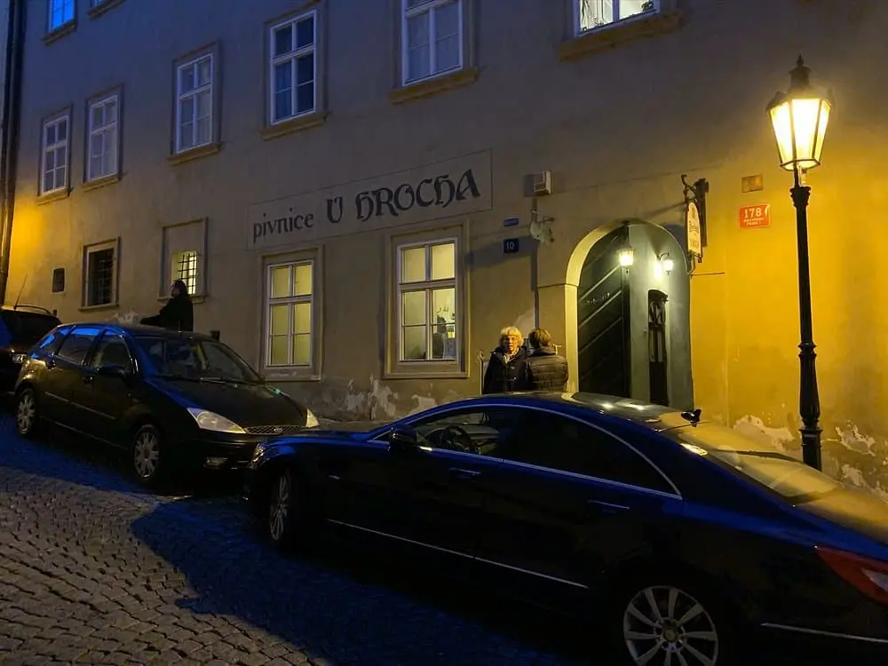 exterier restaurace u hrocha, při večerních hodínách, před hospodou jsou vidět 2 zaparkovaná auta, a u dveří stojí 2 lidé